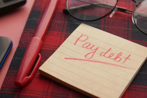 a note that says pay debt next to a pen and glasses by Towfiqu barbhuiya courtesy of Unsplash.