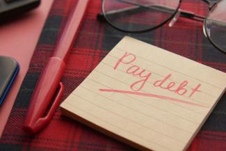 a note that says pay debt next to a pen and glasses by Towfiqu barbhuiya courtesy of Unsplash.